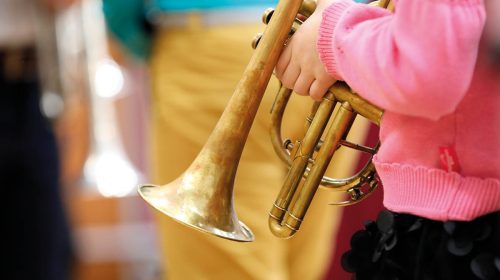 A girl child musician in a pink sweater holds a musical wind instrument trumpet in her hands on a blurred background of an adult teacher.The concept of school music education