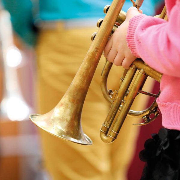A girl child musician in a pink sweater holds a musical wind instrument trumpet in her hands on a blurred background of an adult teacher.The concept of school music education
